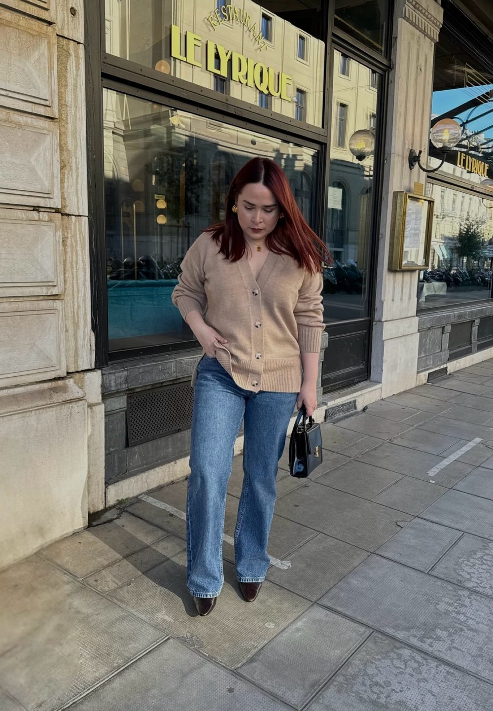 Cardigan beige avec des boutons, jeans évasés bleu clair, sac à main noir et chaussures à talons rouges foncés, le tout sur fond de restaurant.