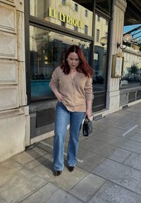 Cardigan beige avec des boutons, jeans évasés bleu clair, sac à main noir et chaussures à talons rouges foncés, le tout sur fond de restaurant.