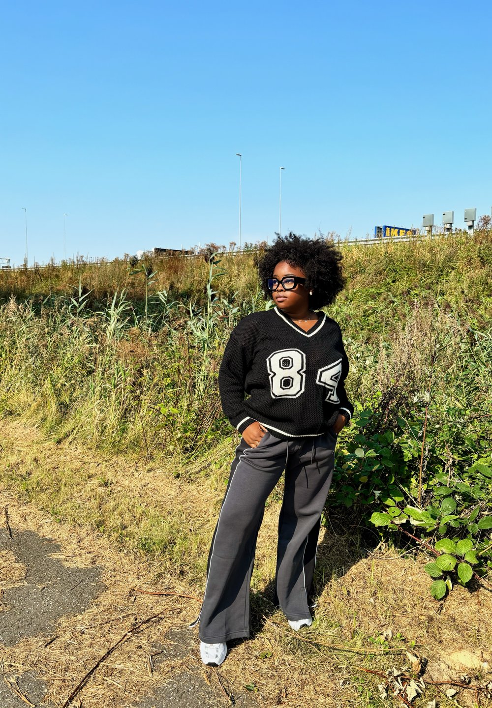 Melna adīta džemperis ar baltām akcentiem un "84" apdruku, saskaņots ar pelēkām platām biksēm un baltām kedām. Dabiska vide ar zaļumiem.