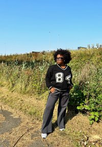 Melna adīta džemperis ar baltām akcentiem un "84" apdruku, saskaņots ar pelēkām platām biksēm un baltām kedām. Dabiska vide ar zaļumiem.