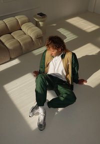 Noor inimene rohelises treeningkostüümis ja spordijalatsites istub päikesepaisteline põrandal, beige diivani ja väikese ümmarguse laua kõrval, millel on raamatud.