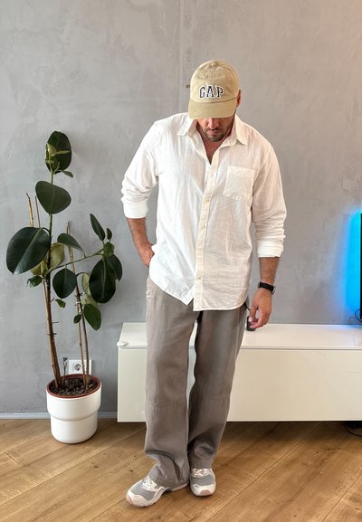 Camisa de lino blanca ligera, gorra beige con el logo de "GAP", pantalones gris claro y zapatillas grises con acentos rosas, sobre un suelo de madera. Planta en maceta cercana.