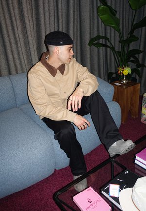 Chaqueta beige con cuello marrón, pantalones negros, zapatos grises. Sentado en un sofá azul. Cortinas oscuras y una planta en el fondo.