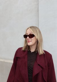 Hnedé slnečné okuliare na osobe, ktorá má na sebe bordový kabát zo semišu a tmavý, textúrovaný top. V pozadí je svetlosivá stena.