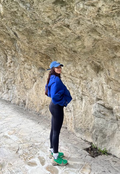 Sudadera azul de gran tamaño, leggings negros, zapatillas verdes y beige, de pie contra un fondo rocoso en un camino de piedra.