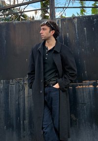 Νεαρός άνδρας με σγουρά μαλλιά φορώντας μακρύ μαύρο παλτό και σκούρα τζιν, στέκεται δίπλα σε έναν φθαρμένο μαύρο μεταλλικό φράχτη, κοιτάζοντας πλάγια.