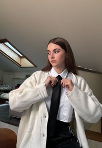 Hvit strikket cardigan over en hvit skjorte med krage, justerer et svart slips, med mørke bukser og et lyst rom med takvindu.