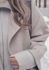Femme portant un manteau en laine beige clair et un pull côtelé crème, tenant le manteau près d'elle, avec des cheveux ondulés lâches visibles sur l'épaule en extérieur.