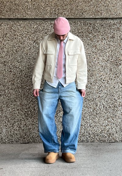 Chaqueta de piel sintética crema, jeans oversized de un azul claro, camisa blanca, corbata rosa y un gorro de punto rosa. Zuecos marrón claro. Fondo texturizado.