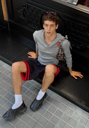 Joven sentado en una acera, vistiendo una camiseta de manga larga gris, pantalones cortos negros con rayas rojas y zapatos negros sin cordones.