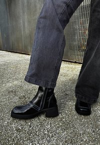 Čierne kožené členkové topánky so štvorcovou špičkou a elastickými bočnými panelmi, s kontrastným modrým prešívaním a masívnym podpätkom. Nosené s šedými džínsami.