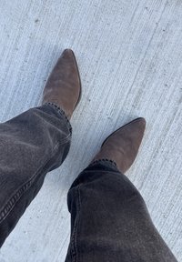 Hnedé semišové čižmy so špicatou špičkou nosené s tmavými džínsami, ukazujúce hladkú textúru a ostrý dizajn na textúrovanom betónovom povrchu.