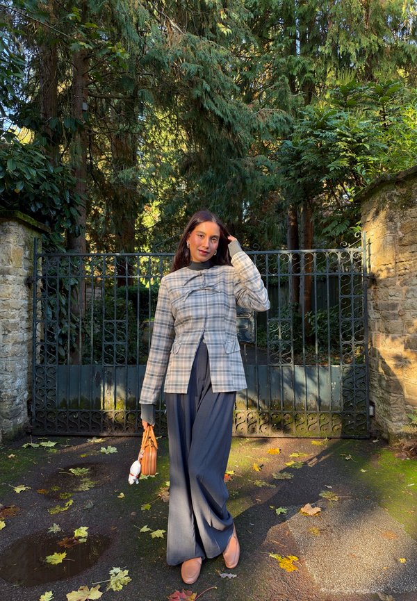 Une femme portant une veste à carreaux et un pantalon à jambes larges pose avec un petit sac à main devant un portail de jardin orné, au milieu des arbres et des feuilles mortes.