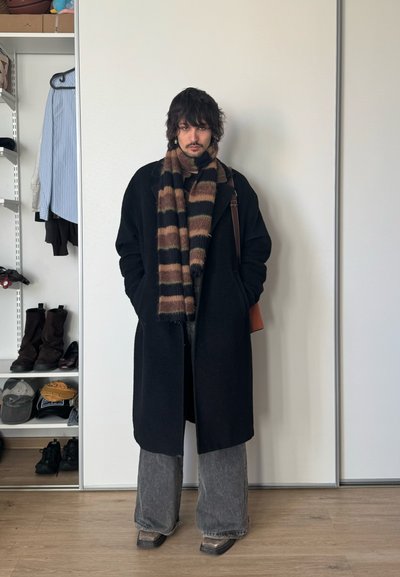 Persona con cabello oscuro que lleva un abrigo negro, una bufanda a rayas marrón y negra, pantalones anchos grises, de pie en el interior cerca de un armario con zapatos y ropa.