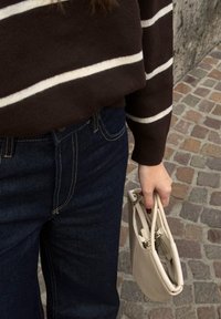 Personne portant un jean bleu foncé et un pull marron avec des rayures blanches, tenant un petit sac à main beige dans une rue pavée.