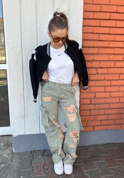 Chaqueta de béisbol negra con rayas blancas, camiseta blanca de Nike, pantalones cargo ligeros verde claro desgastados con desgastes, y zapatillas blancas.