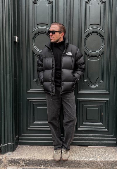 Hombre con chaqueta acolchada negra, cuello alto negro, vaqueros oscuros y gafas de sol, de pie con las manos en los bolsillos frente a una puerta con paneles oscuros.
