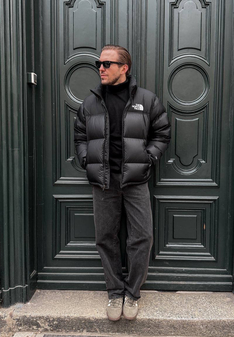 Homme portant une doudoune noire, un col roulé noir, un jean foncé et des lunettes de soleil, debout les mains dans les poches contre une porte à panneaux sombre.