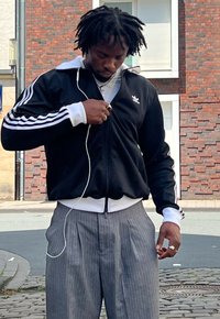 Chaqueta deportiva negra con cremallera y rayas blancas, y un cuello blanco. Pantalones grises a rayas finas, llevando una cadena de plata y anillos.
