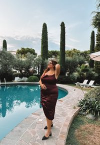 Burgunderfarbenes Satin-Kleid mit gerafften Details, getragen mit schwarzen spitzen Fersen. Umgeben von einem Pool, einem Steinweg und üppigem Grün.
