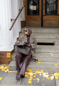 Kraciasty brązowy płaszcz, bordowe skórzane spodnie i brązowe buty. Trzyma szarą torbę z złotym logo. Szare kamienne schody z żółtymi liśćmi.