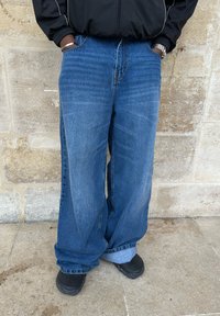 Szerokie niebieskie dżinsy z wyblakłym wykończeniem, o luźnym kroju, z podwiniętymi mankietami, zestawione z czarnym obuwiem.