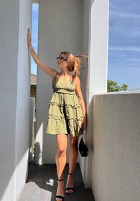 Femme portant des lunettes de soleil et une robe vert olive posant sur un balcon baigné de soleil, tenant un sac à main noir et touchant un mur blanc.