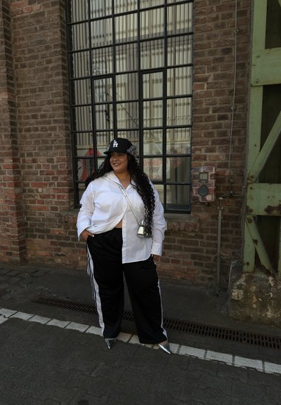 Camisa blanca de botones, pantalones deportivos negros con rayas blancas, zapatos puntiagudos plateados y una gorra negra. Cabello rizado y una pequeña bolsa cruzada.