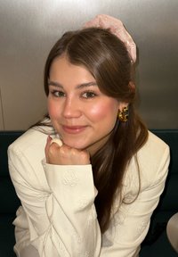 Kremowa marynarka z kwiatowym haftem, złote kolczyki koła, przezroczysta różowa gumka do włosów i kremowy pierścionek, pozująca zrelaksowanym wyrazem twarzy.