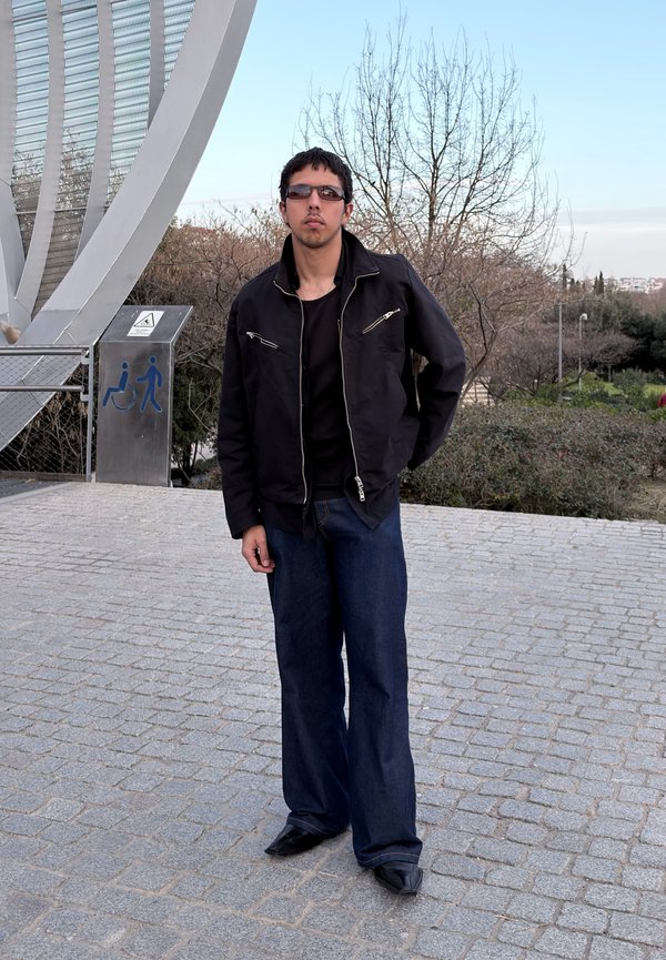 Homme portant une veste foncée, des lunettes de soleil et un jean large, debout sur une zone pavée près d'une structure métallique et d'un panneau d'accessibilité en extérieur.