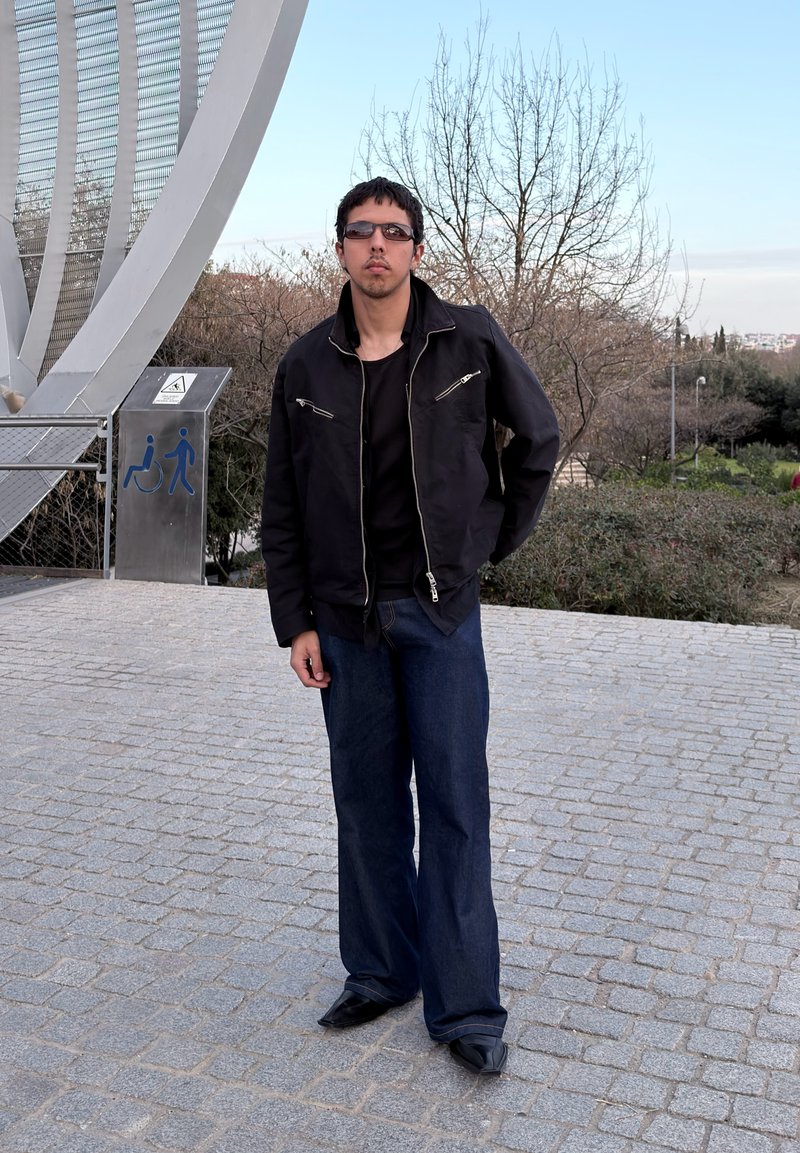 Άνδρας φοράει σκούρο μπουφάν, γυαλιά ηλίου και φαρδιές τζιν μπλούζες, στέκεται σε πλακόστρωτη περιοχή κοντά σε μεταλλική κατασκευή και πινακίδα προσβασιμότητας σε εξωτερικό χώρο.