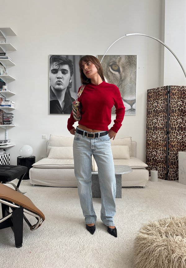 Mujer con suéter rojo y vaqueros azul claro está en una sala de estar moderna con sofá crema, un gran león, retratos en blanco y negro y un biombo con estampado de leopardo.