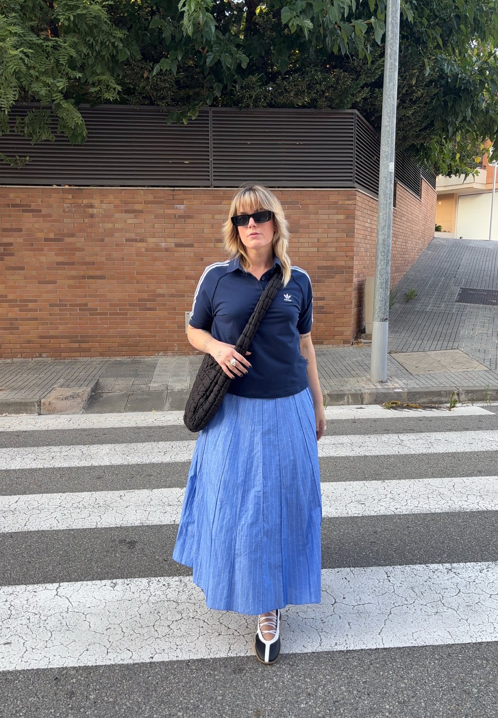 Polo de cor azul marinho com riscas brancas, saia maxi plissada azul, saco de fibras pretas e sapatos de salto alto brancos e pretos numa passadeira.