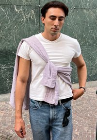T-shirt bianco a maniche corte indossato con una camicia a quadri chiara legata davanti. I jeans blu e occhiali da sole neri pendono dalla cintura.