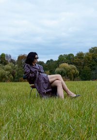 Barna bőrcsuklya, fekete napszemüveg, egy széken ül egy fűvel borított mezőn, mögötte fák és felhős égbolt.