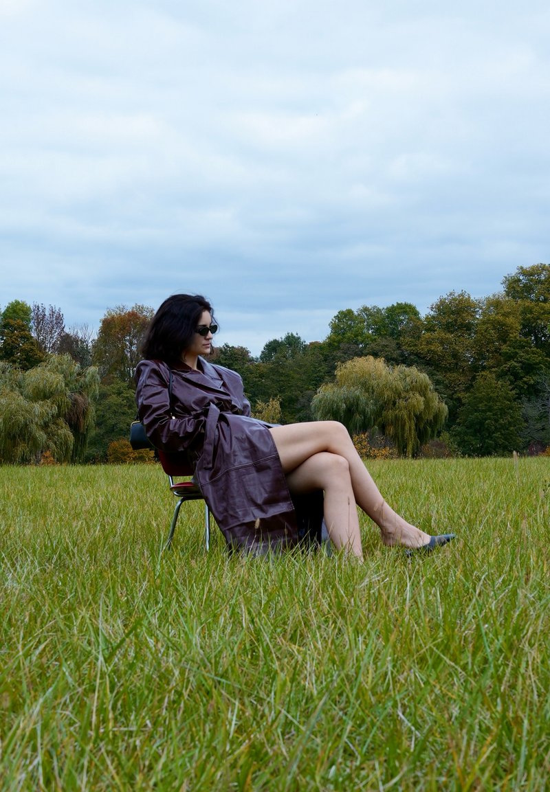 Bruin leren trenchcoat, zwarte zonnebril, zittend op een stoel in een grasveld met op de achtergrond bomen en een bewolkte lucht.