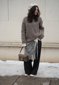 Femme portant un béret beige, un pull marron pelucheux avec des détails en dentelle, un pantalon large noir et des chaussures crème, debout sur un trottoir enneigé tenant un sac à main taupe.