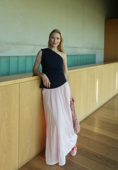 Top azul marino de un solo hombro combinado con una falda larga de capas en rosa. Sosteniendo una bolsa trenzada en rosa claro. Suelo de madera y fondo minimalista.