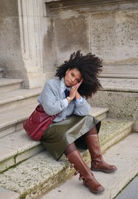 Cardigan grigio a maglia, colletto di pizzo bianco, gonna in vinile verde, stivali marroni alti fino al ginocchio con fibbie e borsa a spalla rossa. Seduta sui gradini di pietra.