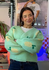 Jeune femme aux cheveux bruns courts, portant des boucles d'oreilles créoles et un pull vert menthe orné de fleurs crème, debout les bras croisés à l'intérieur.