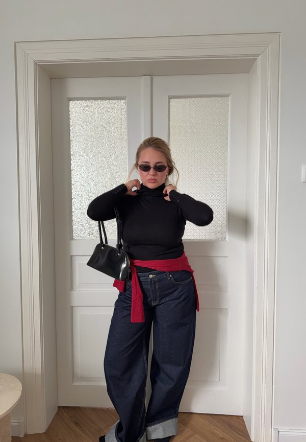 Kobieta w czarnym golfie, ciemnych szerokich dżinsach, czerwonym swetrze zawiązanym w talii, małej czarnej torebce i okularach przeciwsłonecznych, stojąca wewnątrz przy białych drzwiach.
