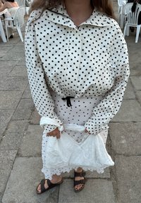 Fehér pöttyös dzseki, deréknál meghúzva, fehér ruha fölött rétegezve. Fehér hímzett táskát tart a kezében; barna szandált visel.