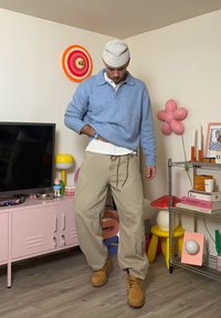 Pull bleu clair en tricot par-dessus une chemise blanche, pantalon ample beige et bottes couleur fauve. La pièce se distingue par une décoration colorée et une unité de rangement rose.