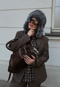 Chapeau d'hiver en fourrure brune, lunettes de soleil surdimensionnées, sac en daim brun foncé avec fermetures éclair argentées, chemise à carreaux sous un blazer marron.