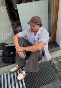Muž sedí na římsě, má na sobě proužkované tričko s krátkým rukávem, hnědou kšiltovku, béžové kalhoty a hnědé a bílé mokasíny, v ruce drží zelený nápoj.