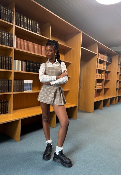 Vestido a cuadros marrón con un corsé ajustado y falda acampanada, combinado con una camisa blanca y zapatos de plataforma negros, con un fondo de biblioteca.