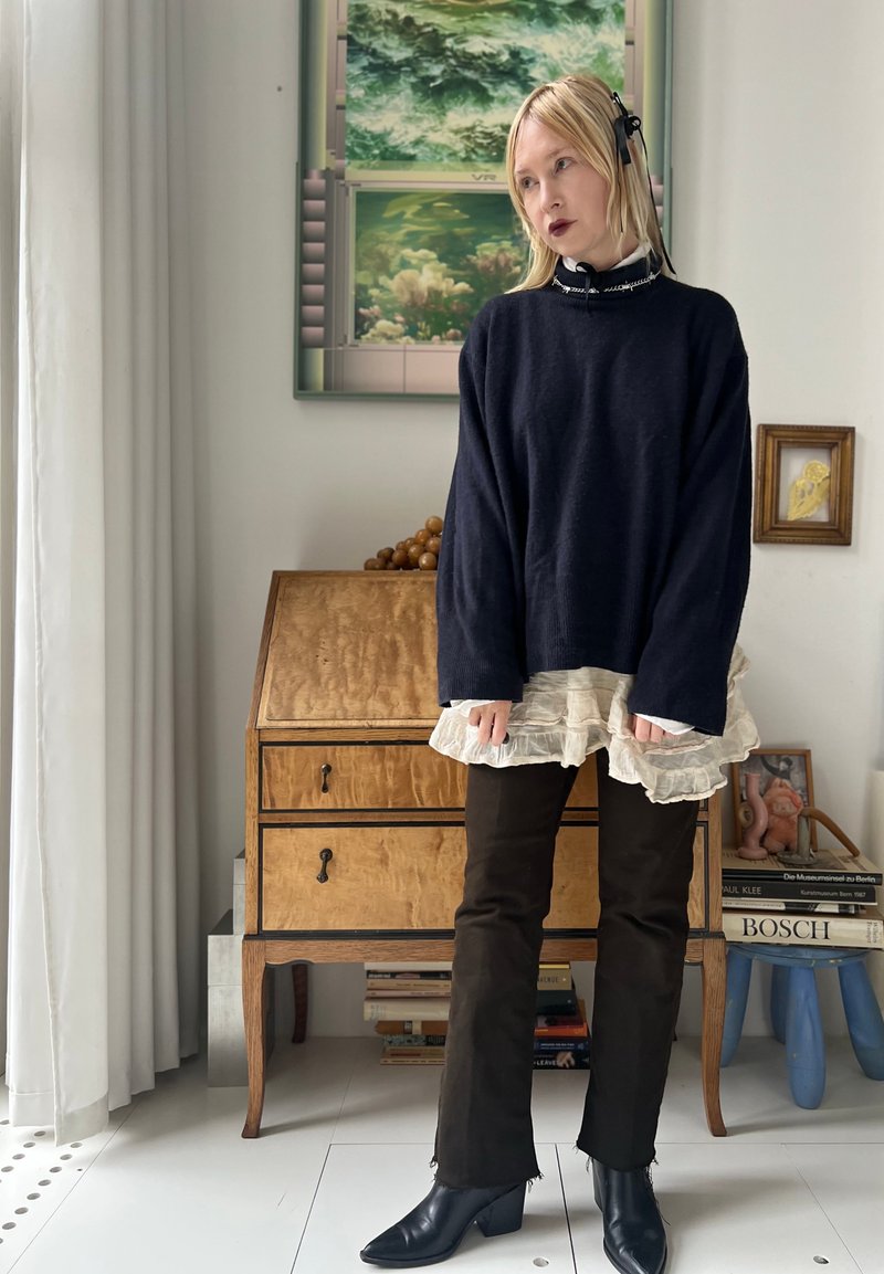 Femme aux cheveux blonds portant un pull noir, une jupe blanche en couches, un pantalon noir et des bottes se tient dans une pièce avec un bureau en bois et des livres.
