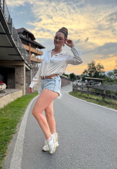 Blusa de satén blanca de manga larga, pantalones cortos de mezclilla claros con bordes deshilachados y zapatillas blancas gruesas. Entorno al aire libre con un fondo pintoresco.