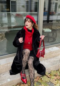Pelliccia sintetica nera, dolcevita rosso, berretto rosso, collant a pizzo, gonna nera, borsa rossa e décolleté rosse in vernice con fibbie.