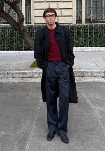 Joven con cabello rizado y gafas, vestido con un abrigo negro, suéter rojo, pantalones azul marino y zapatos negros, de pie en una acera de la ciudad.